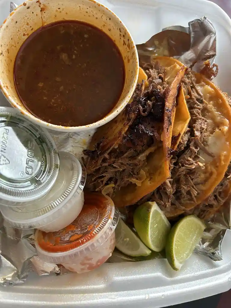 A close-up photo of a takeout container holding two tacos filled with shredded meat, accompanied by a bowl of dark sauce…