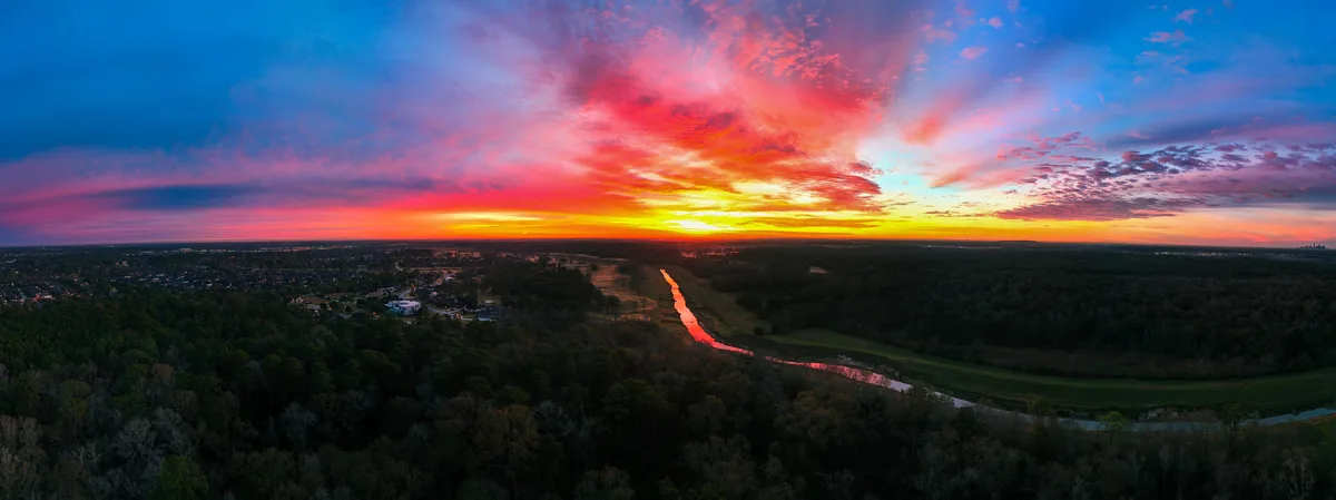 A panoramic aerial photo captures a stunning sunset with vibrant red, orange, and blue clouds over a landscape of forests, a…