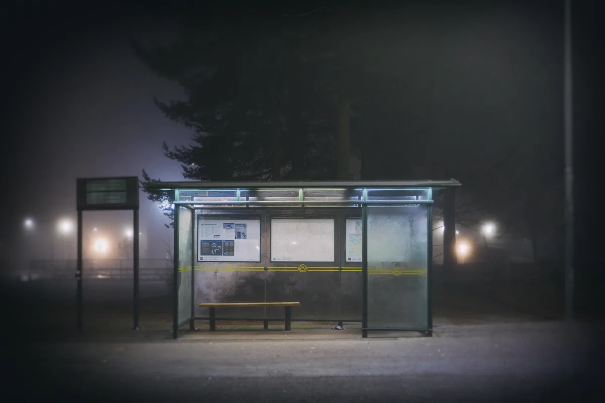 A dimly lit, foggy night scene featuring an empty bus stop with a wooden bench, glass panels, and route maps. The overlaid…