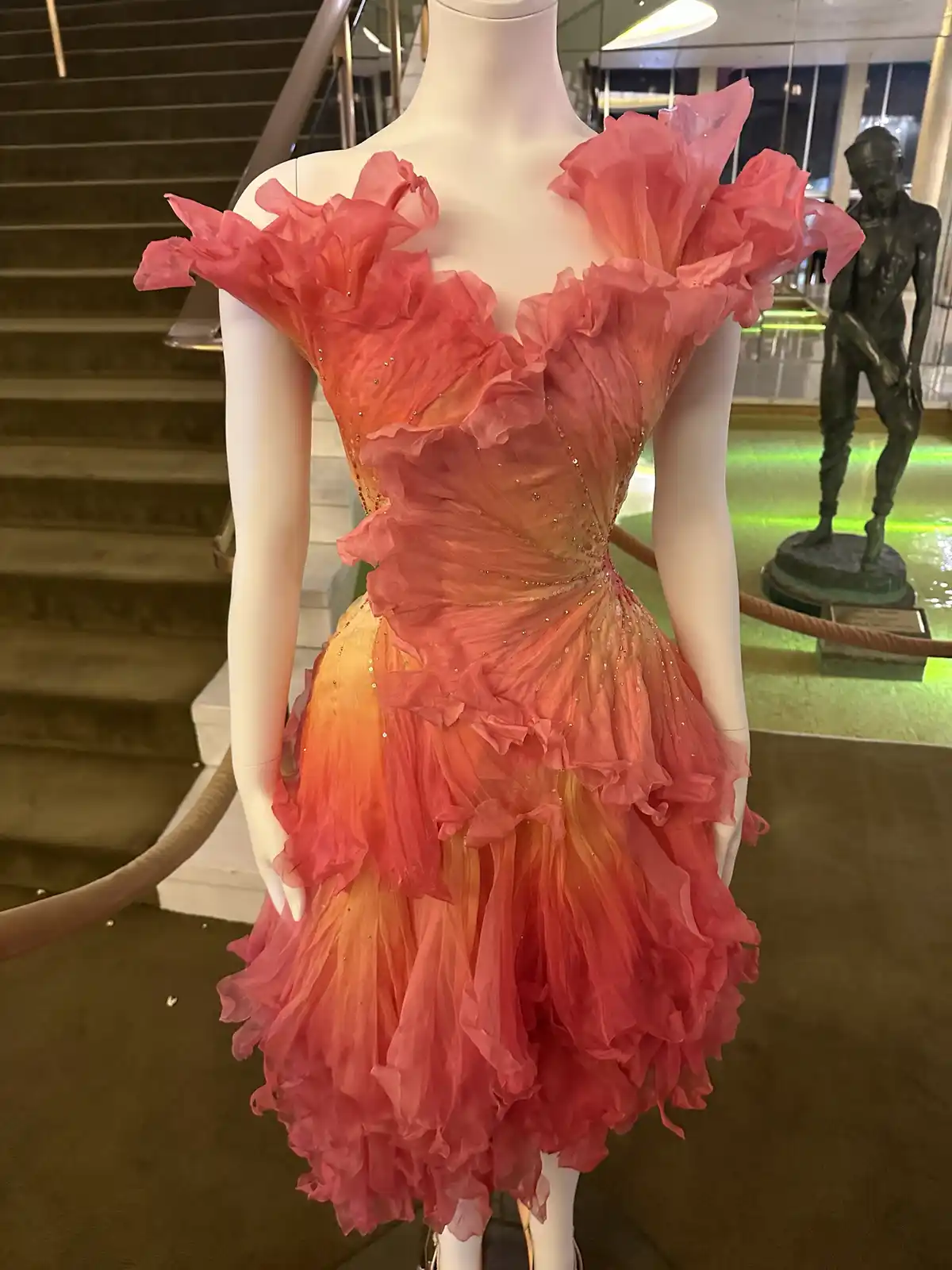 A white mannequin displays a vibrant, ruffled dress with a gradient of pink to orange, adorned with sequins. The dress has…