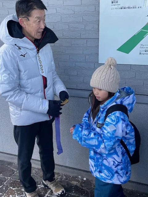 An older man in a white winter jacket holds a medal with a purple ribbon while standing next to a young girl in a blue…