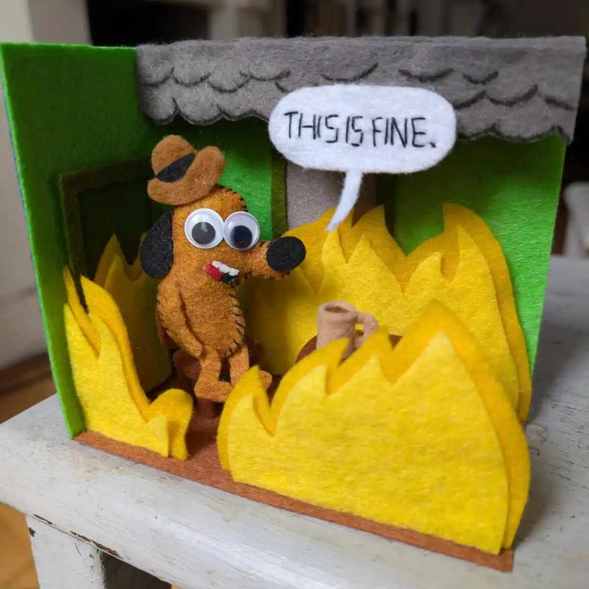 A handmade felt diorama shows a cartoon dog wearing a hat and smoking a pipe, standing calmly inside a house surrounded by…