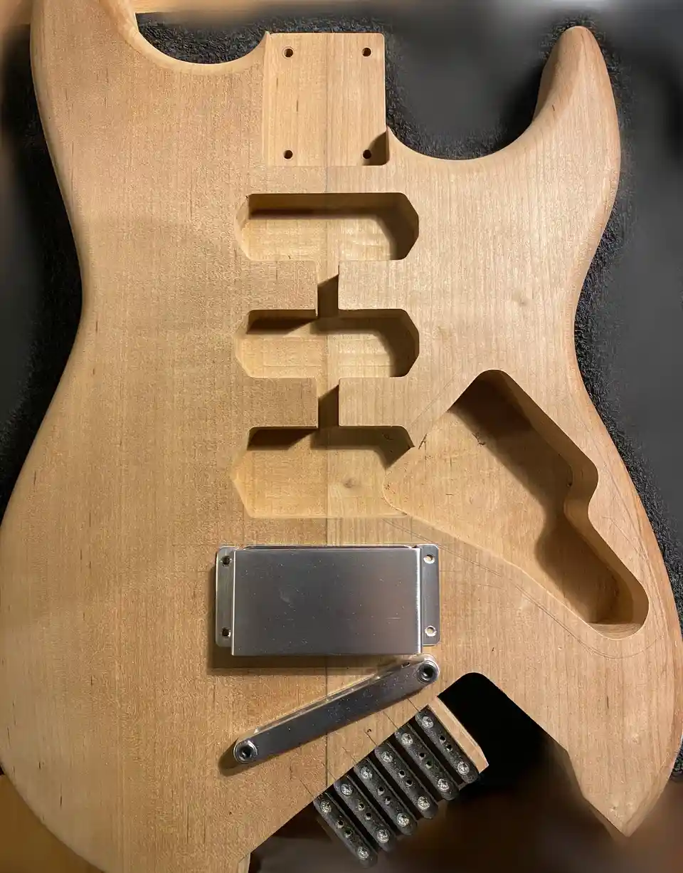 A close-up photo of an unfinished, raw wood electric guitar body with visible cutouts for pickups and bridge, showing the…