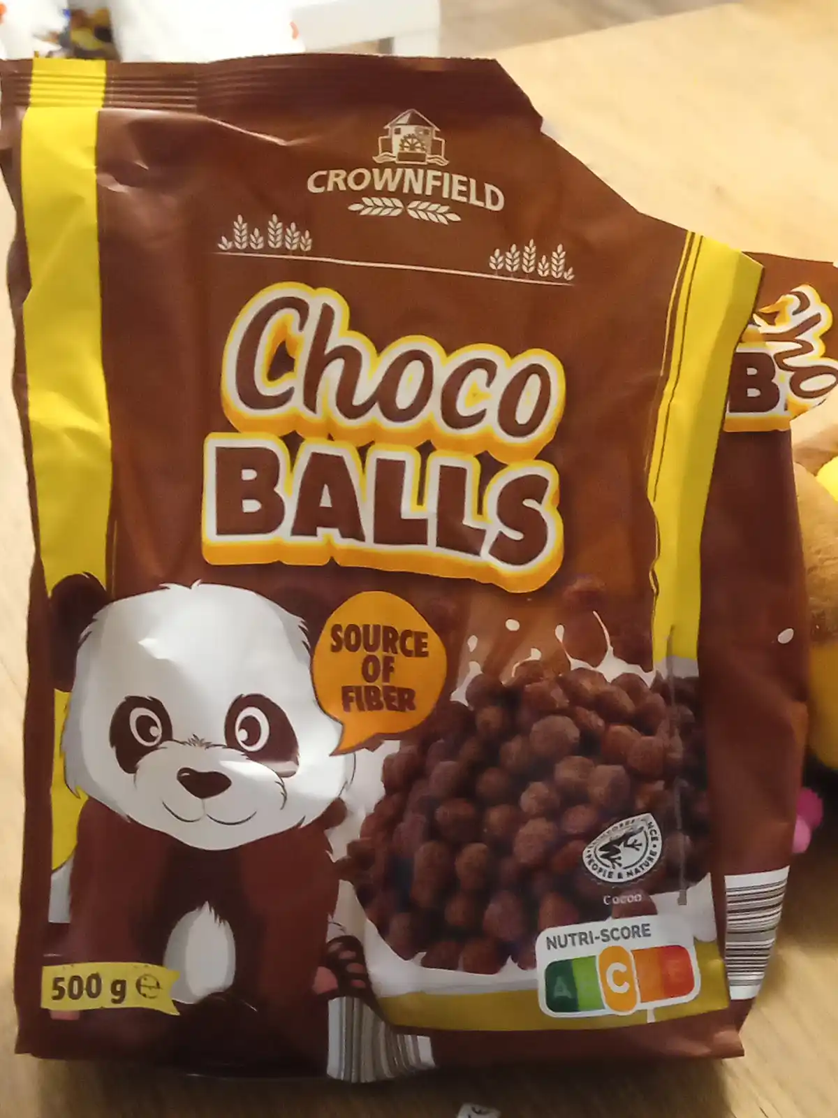 A brown and yellow bag of Crownfield Choco Balls cereal features a cartoon panda holding a speech bubble that says 'source…
