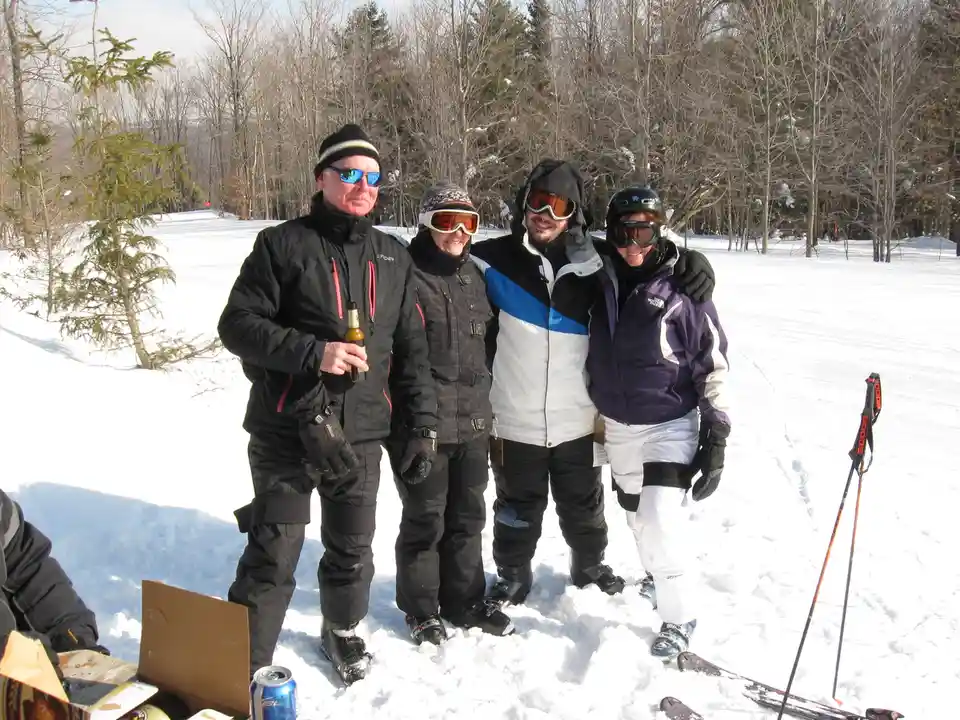 Four people in ski gear stand together on a snowy slope, smiling for a photo. The person on the far left holds a beer…