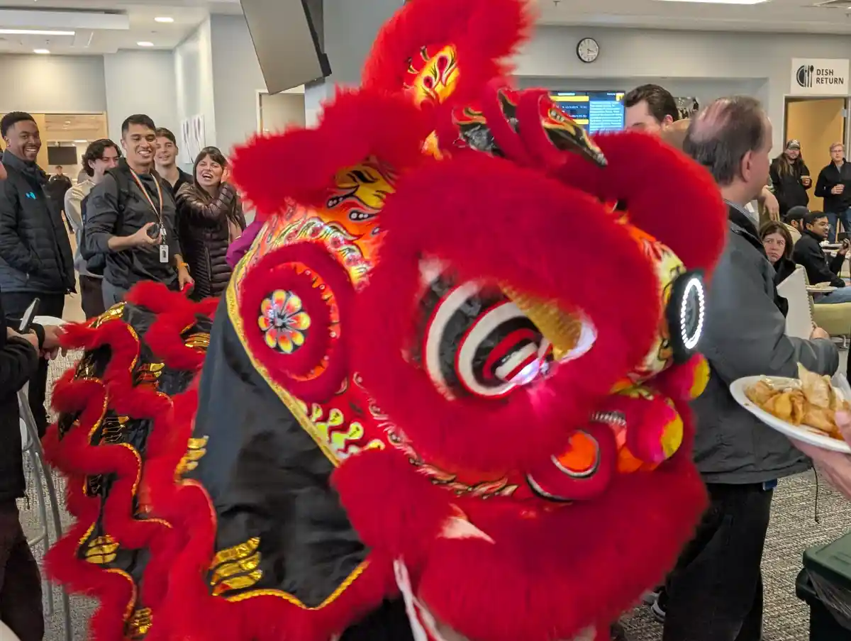 A vibrant red and gold lion dance costume with glowing LED eyes is in the foreground, surrounded by people in a…