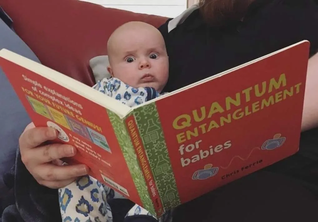 A baby with wide eyes is being held while looking at a red book titled 'QUANTUM ENTANGLEMENT for babies' by Chris Ferrie. An…