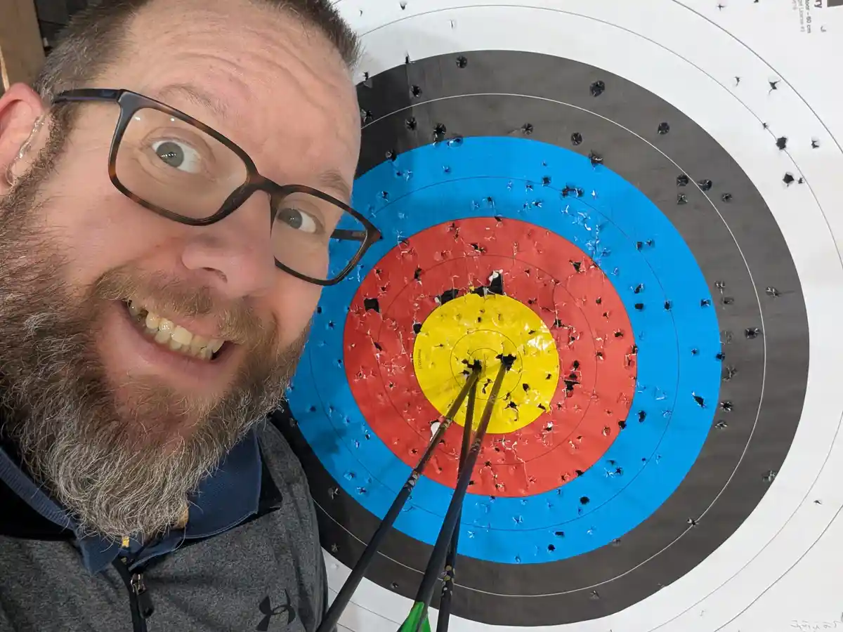 A man with glasses and a beard smiles widely at the camera while standing beside a heavily damaged archery target. The…