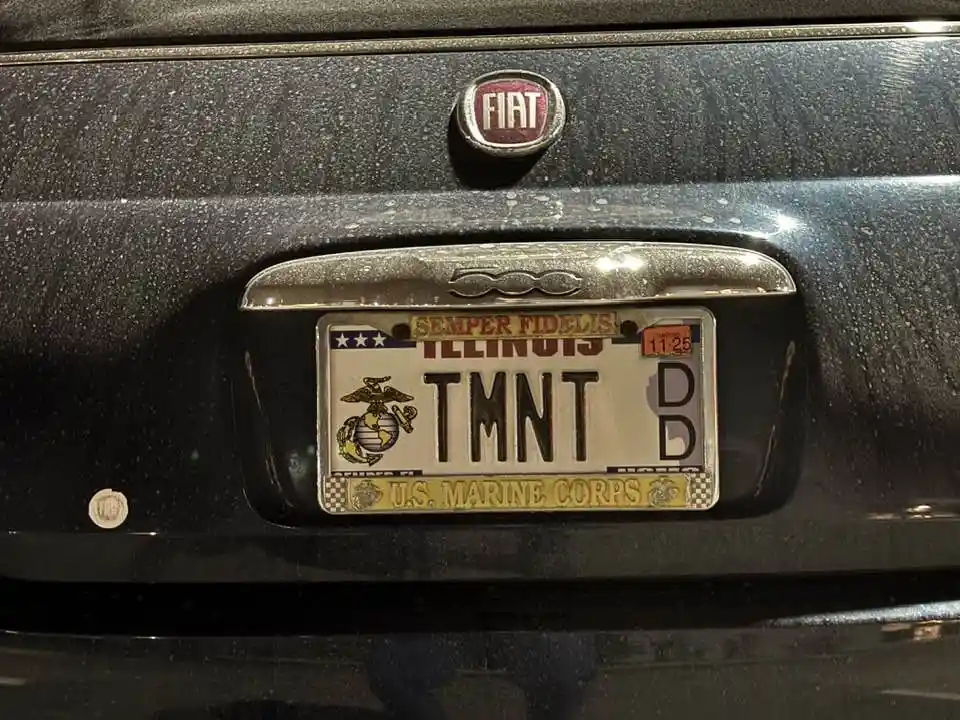 A close-up of a black Fiat car's rear shows an Illinois license plate with the letters 'TMNT' and a U.S. Marine Corps theme…