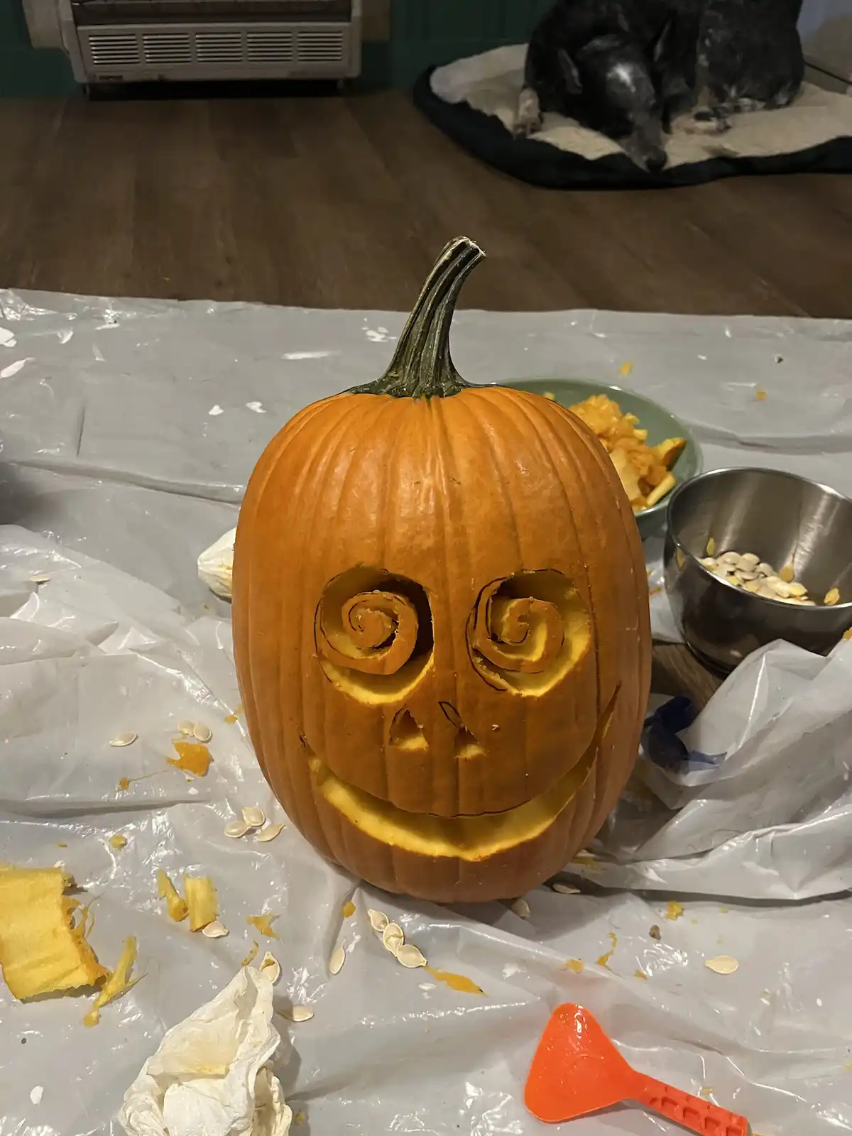 A carved orange pumpkin with spiral eyes and a wide grin sits on a plastic sheet, surrounded by pumpkin guts, seeds, and…