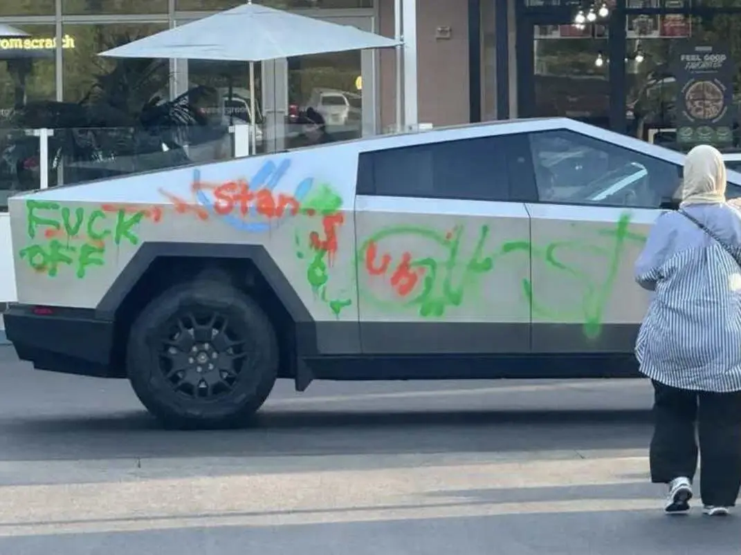 A white Tesla Cybertruck is covered in colorful graffiti, including the words 'FUCK OFF' in green spray paint on its rear. A…