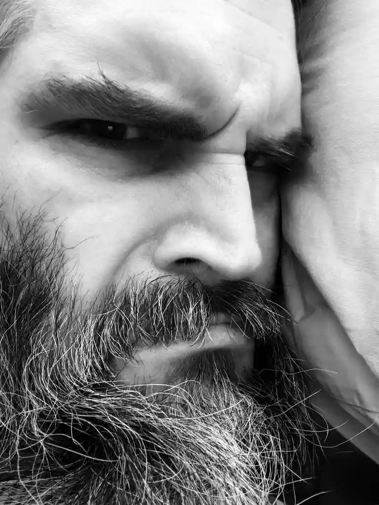 A black-and-white close-up of a man with a thick, graying beard and mustache, his face scrunched in an expression of intense…
