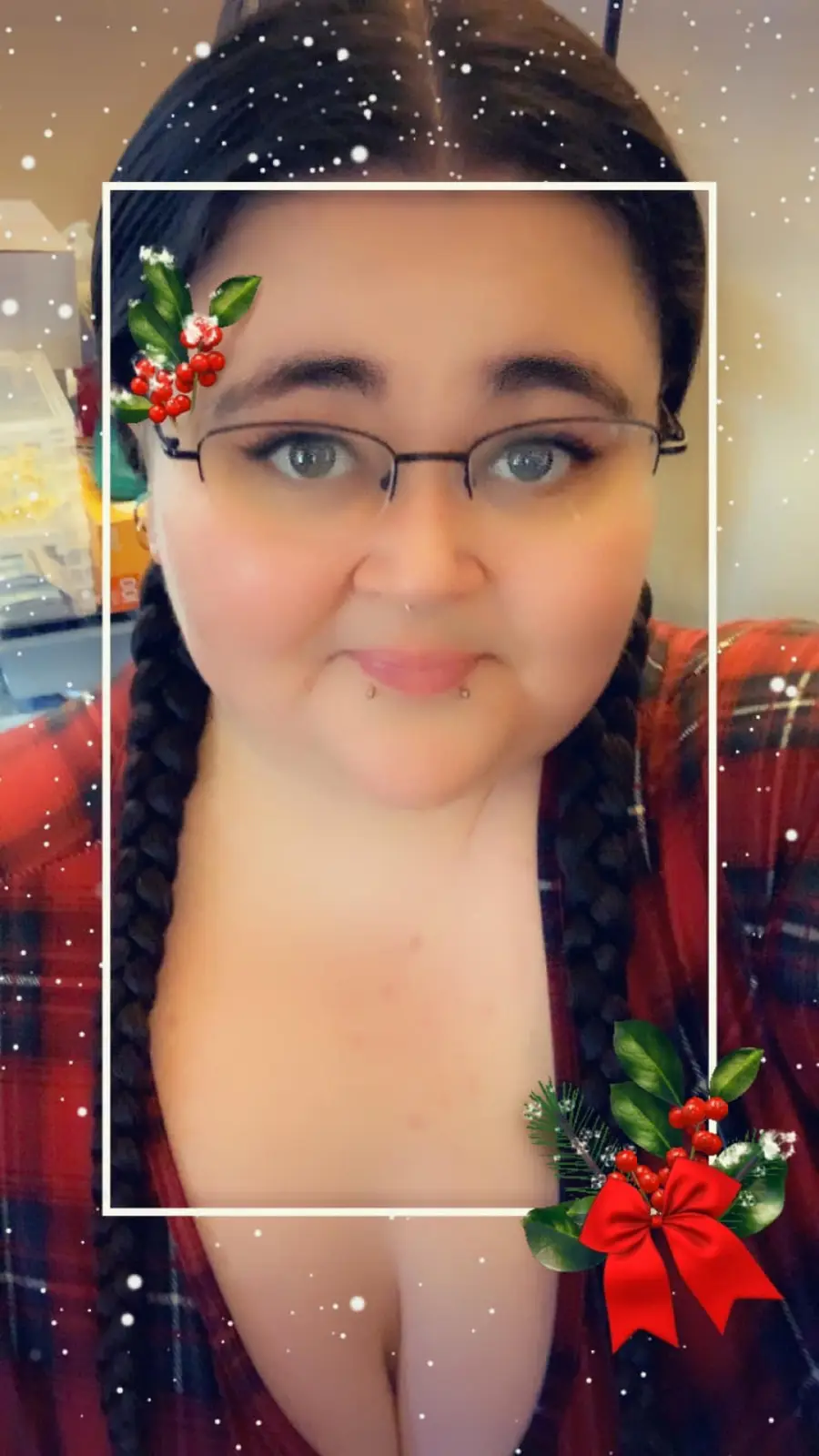 A selfie of a person with dark braided hair, glasses, and a red plaid shirt, framed with a white border and festive holly…