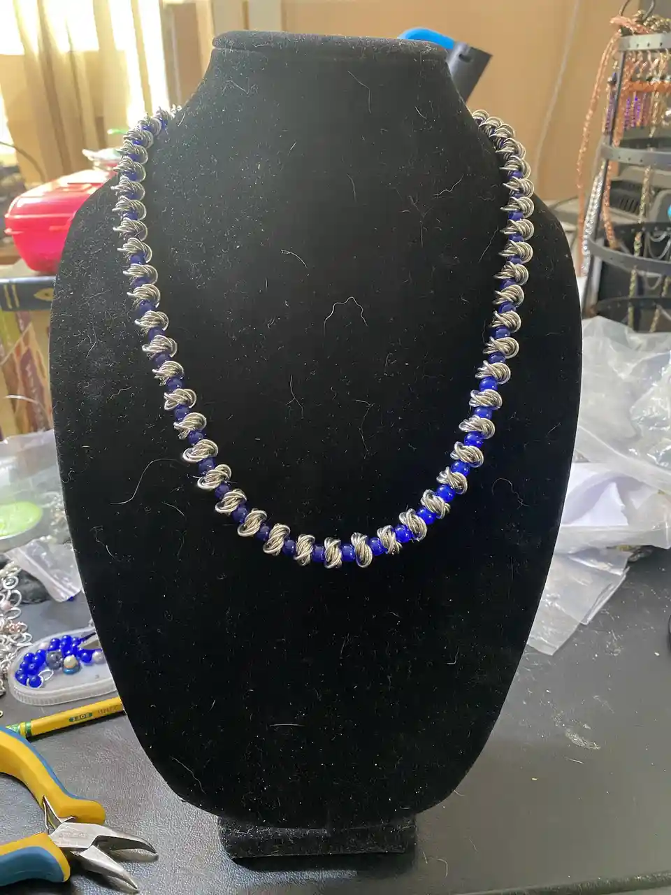 A handmade necklace with blue beads and silver wire wrapping is displayed on a black velvet jewelry bust. The background…