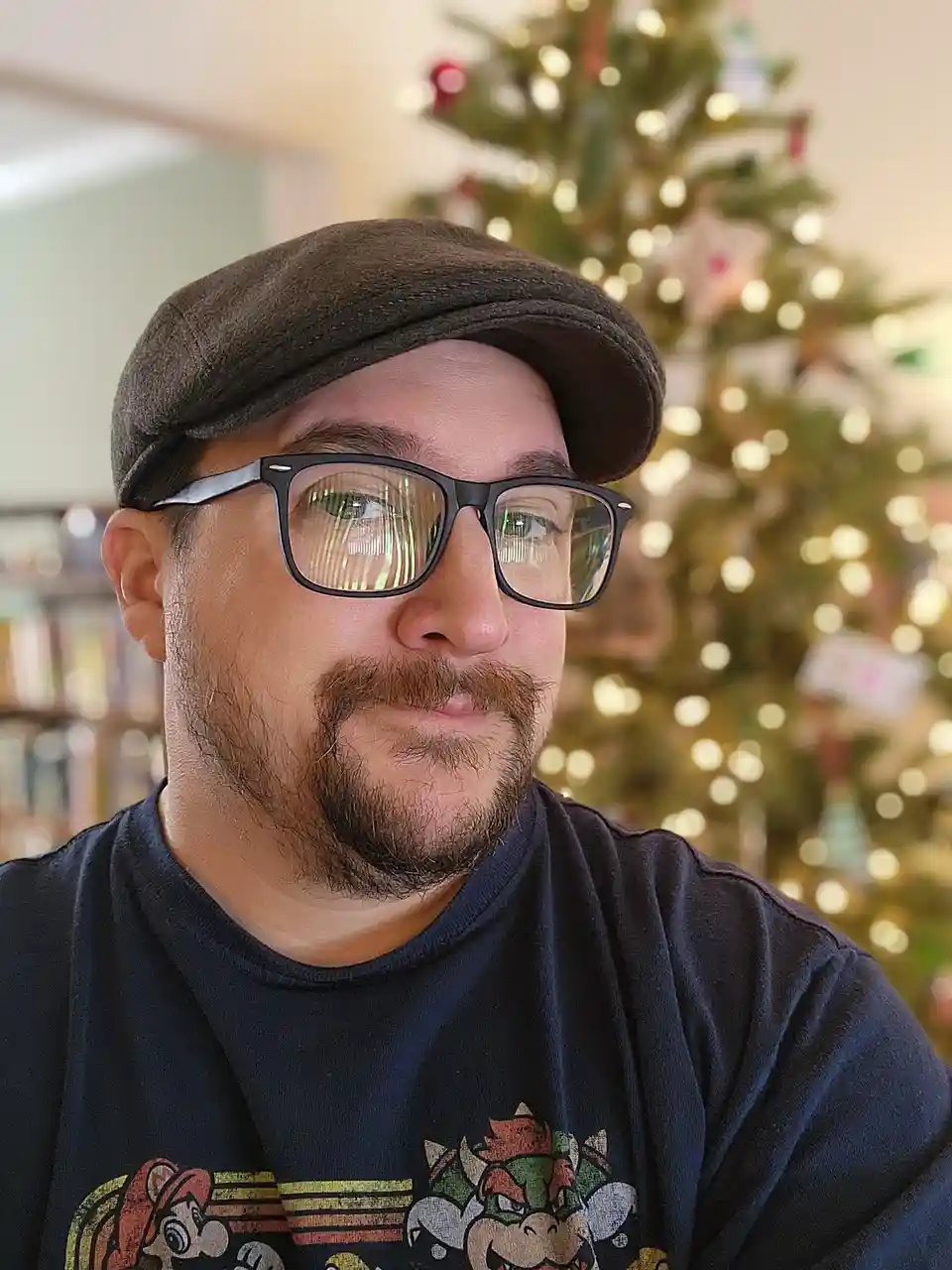 A man with glasses and a flat cap looks directly at the camera with a slightly smug or unimpressed expression. He wears a…