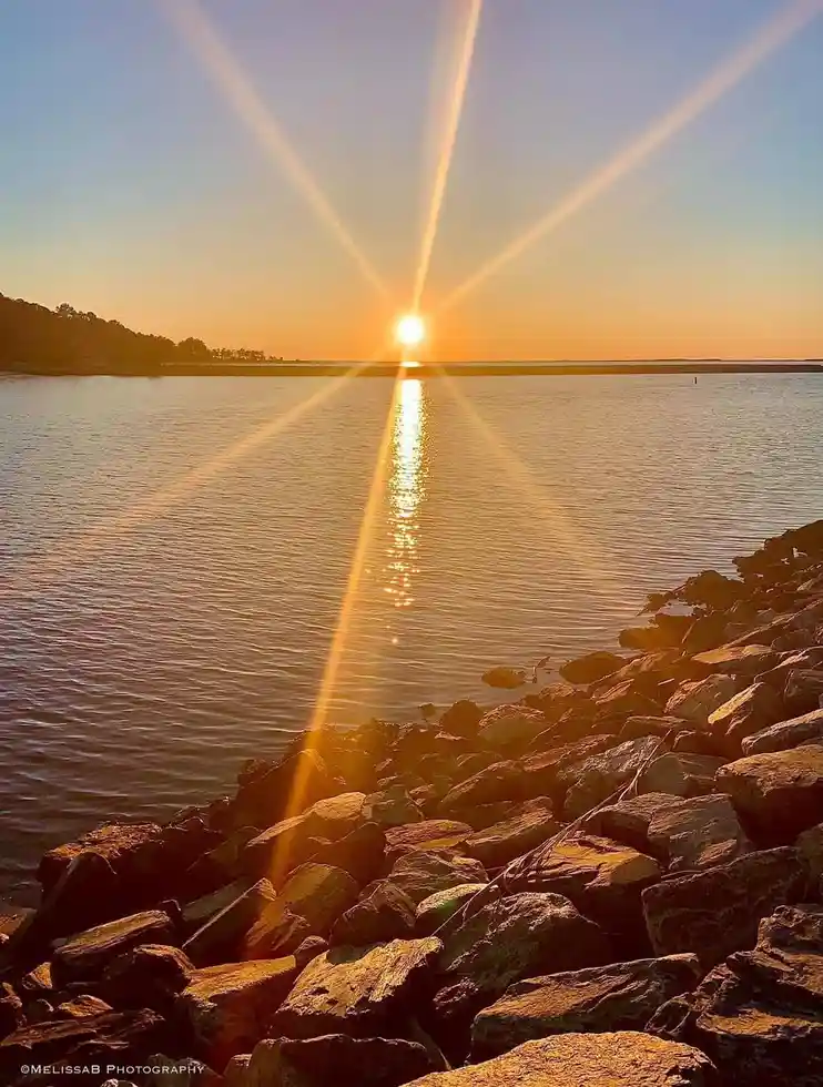 A scenic photo of a sunset over water with a lens flare effect, featuring a rocky shoreline in the foreground. The sun is…