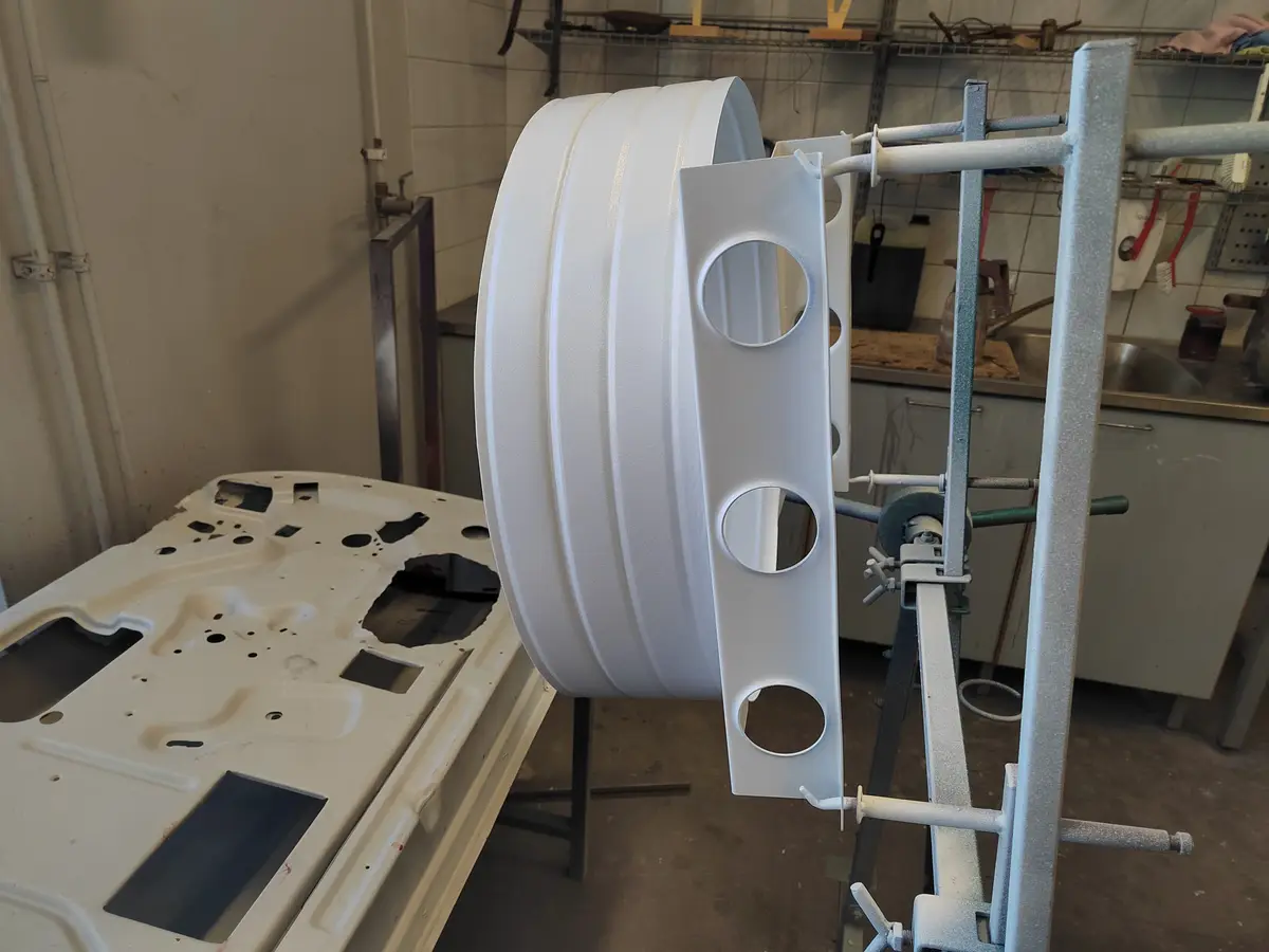 A white-painted car part, possibly a fender or wheel arch, is mounted on a metal stand in a workshop. Beside it, another car…