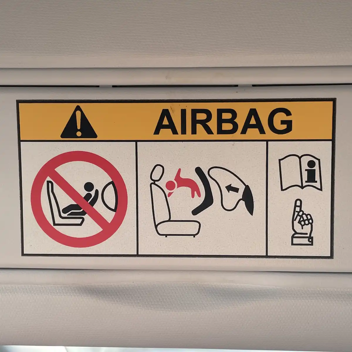 A close-up of a car's airbag warning label showing three panels: left panel has a red circle with a slash over a person…