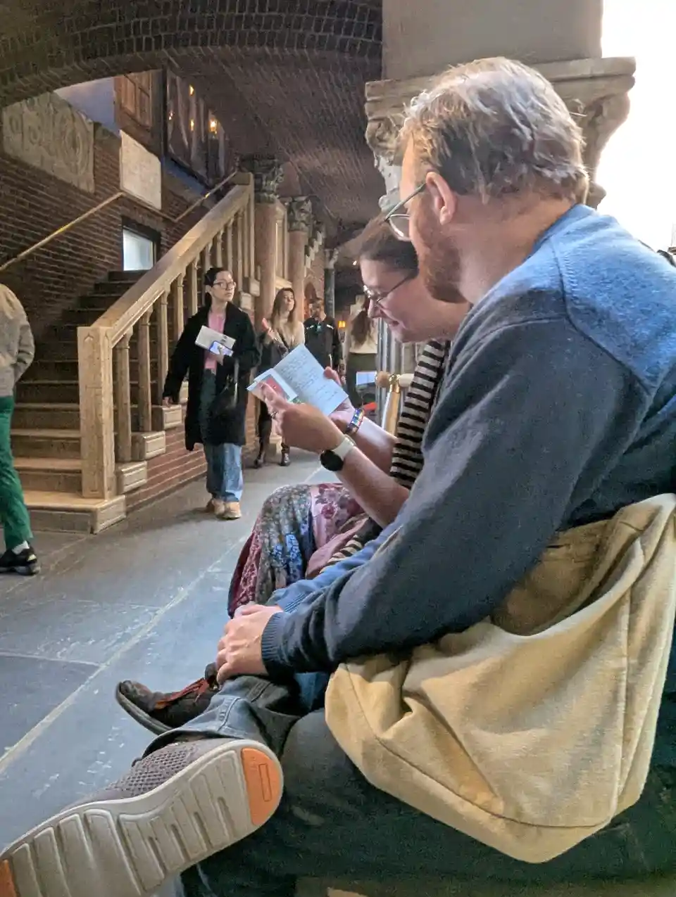 Two people sit on a bench in a historic-looking indoor corridor with brick walls and wooden railings, both looking at paper…