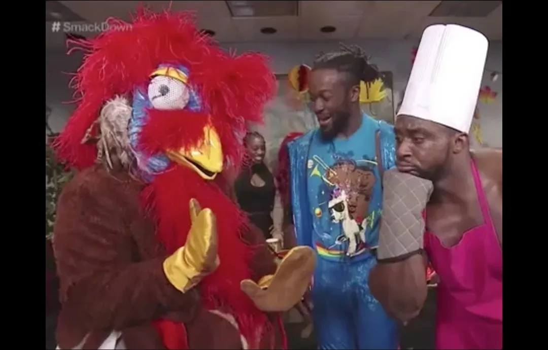 A red-feathered bird mascot in a brown suit and yellow gloves gestures while standing next to a man in a blue sequined…