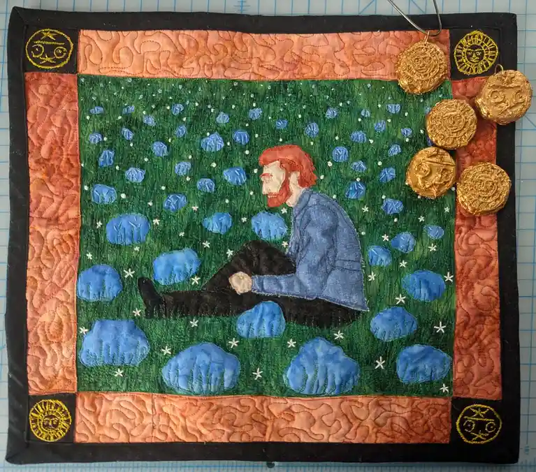 A handcrafted quilt depicts a man with red hair and beard sitting on grass, holding a small golden coin. The quilt features…