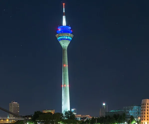 A tall, slender tower illuminated at night with blue and white lights around its upper section and red lights along its…