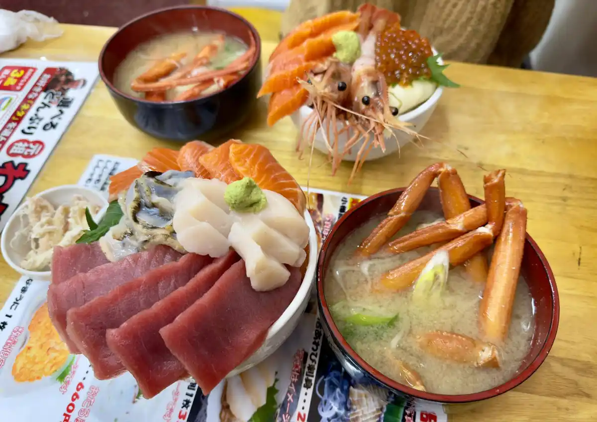 A table is laden with multiple Japanese seafood dishes including sashimi bowls with tuna, salmon, and other fish, a bowl of…