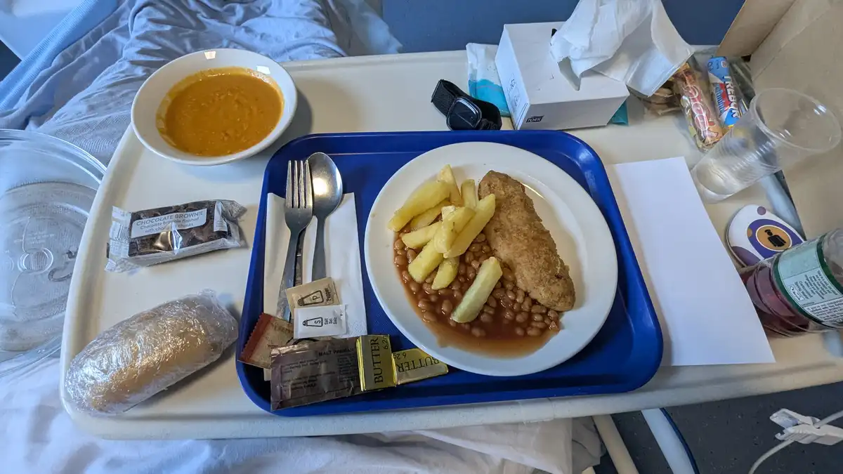 A hospital meal tray with a plate of fish, chips, and baked beans, a bowl of soup, and a chocolate brownie, all set on a…