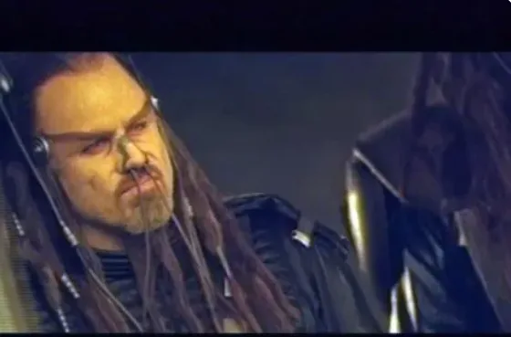 A man with long dreadlocks and a goatee, wearing a dark jacket and headphones, stares intensely with a scowl. The overlaid…