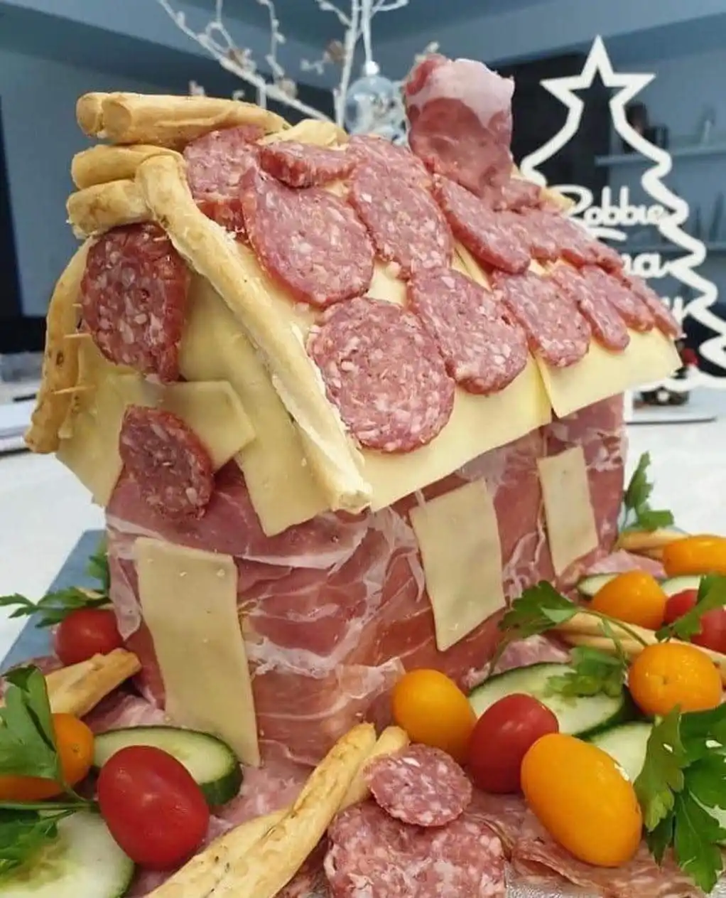 a whimsical food arrangement shaped like a house, constructed from layers of salami slices forming the roof, cheese slices…