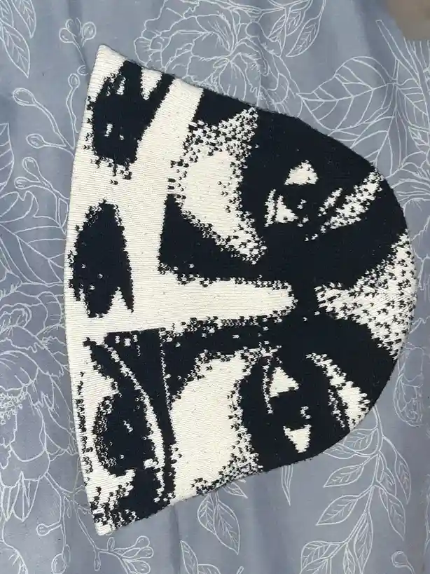 A black and white knitted hat with a pixelated cat face design lies on a blue fabric with white floral patterns. The…