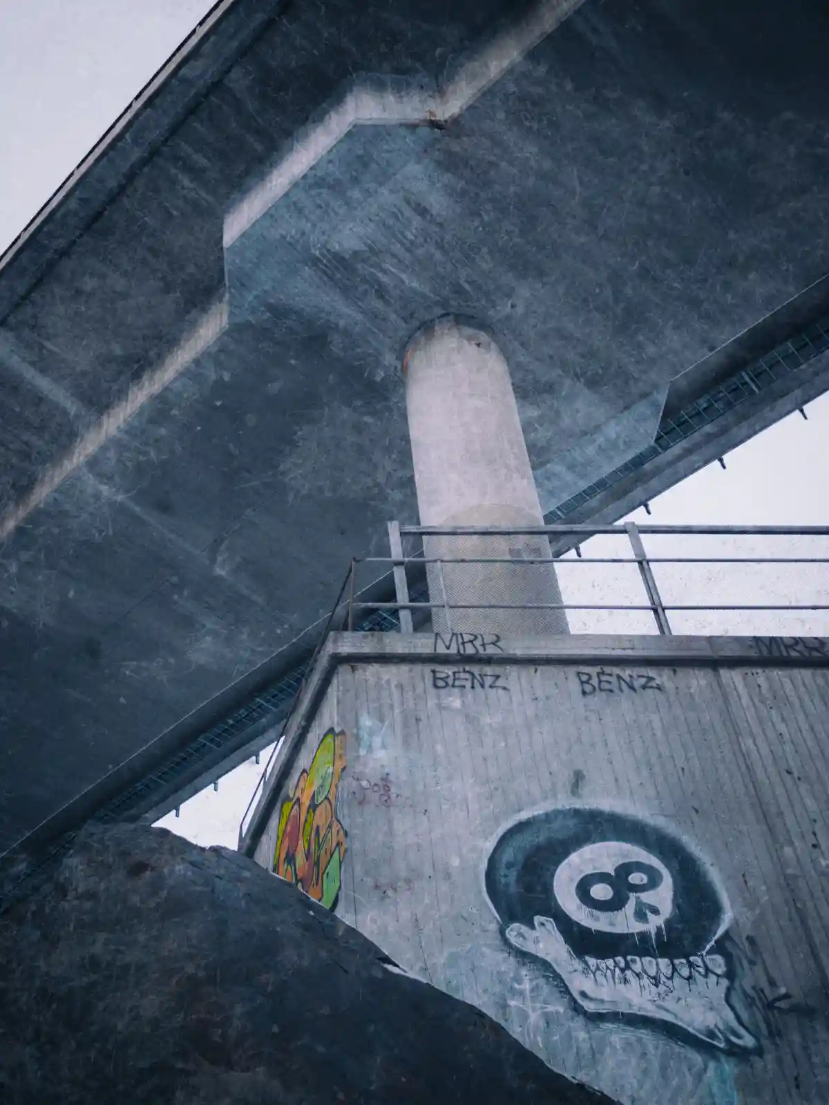 A low-angle shot looks up at the underside of a concrete overpass, showing graffiti on the support pillar including the word…