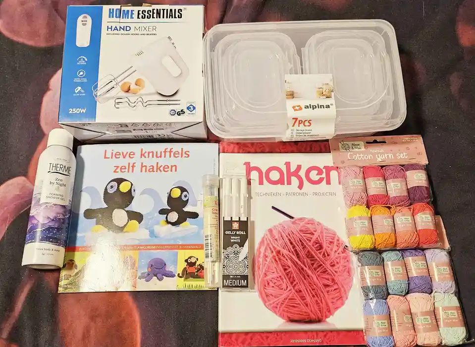 An overhead shot displays a collection of household and craft items including a boxed hand mixer, clear plastic storage…