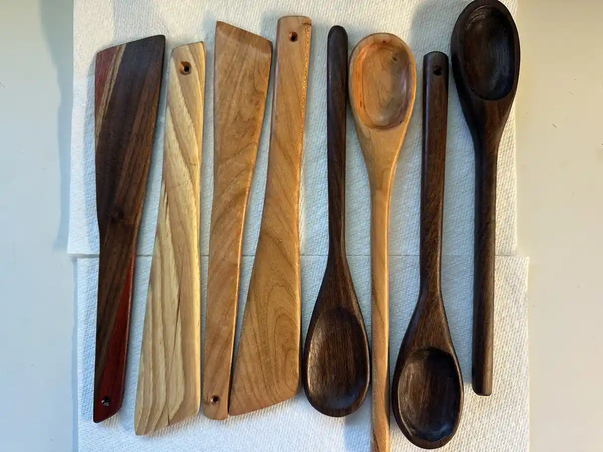 A top-down photo shows eight handcrafted wooden kitchen utensils—four spatulas and four spoons—arranged neatly on a white…