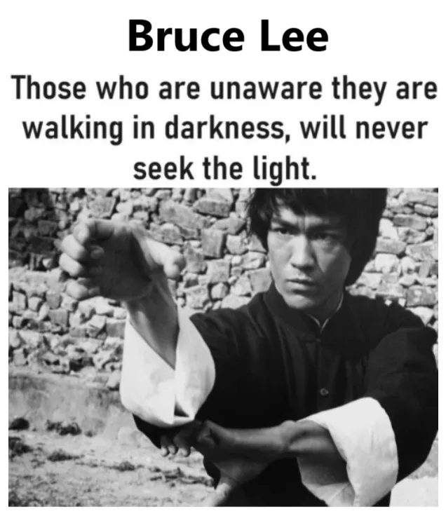 A black and white photo of Bruce Lee in a martial arts stance, wearing a dark shirt with white sleeves, against a stone wall…