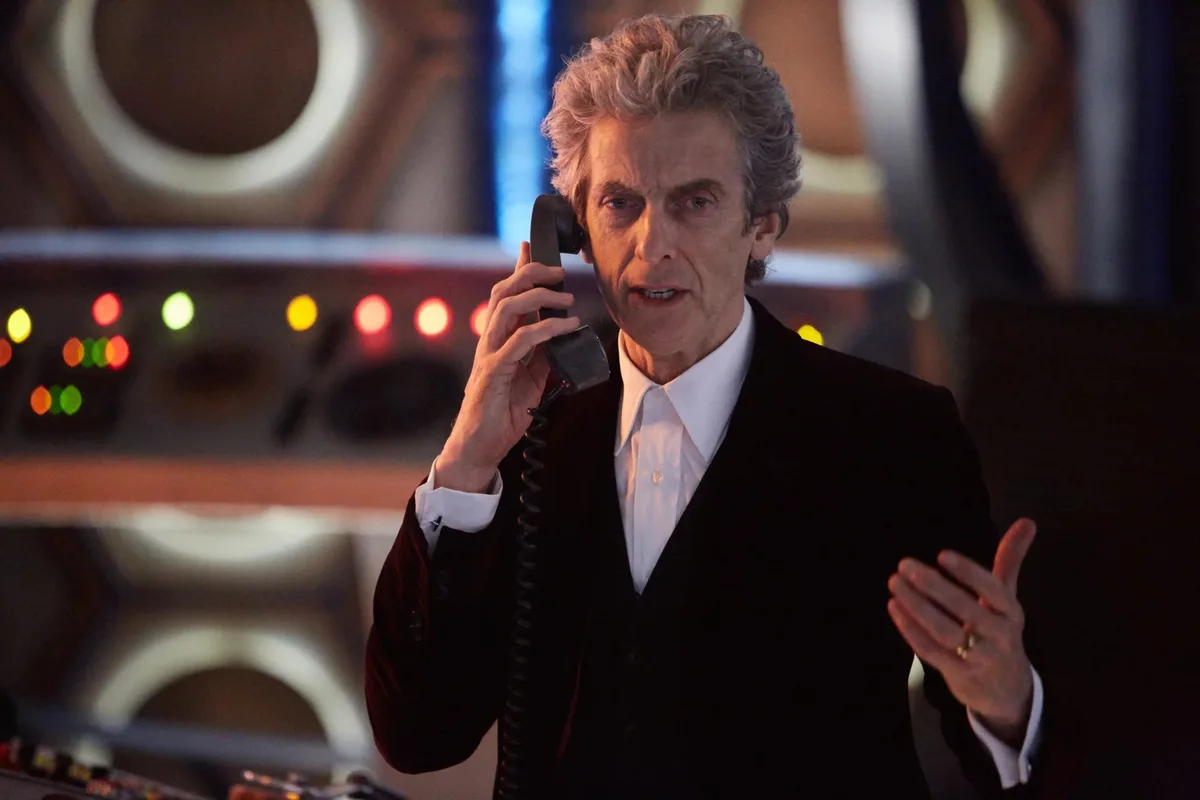 A man with curly gray hair, wearing a black velvet suit and white shirt, holds a vintage telephone receiver to his ear while…