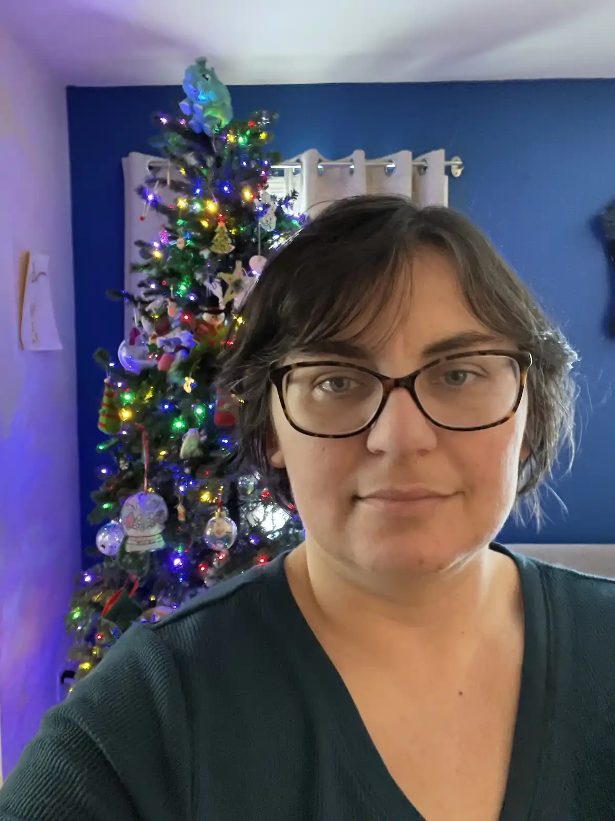 A person with short dark hair and glasses looks at the camera with a neutral expression, standing in front of a brightly…