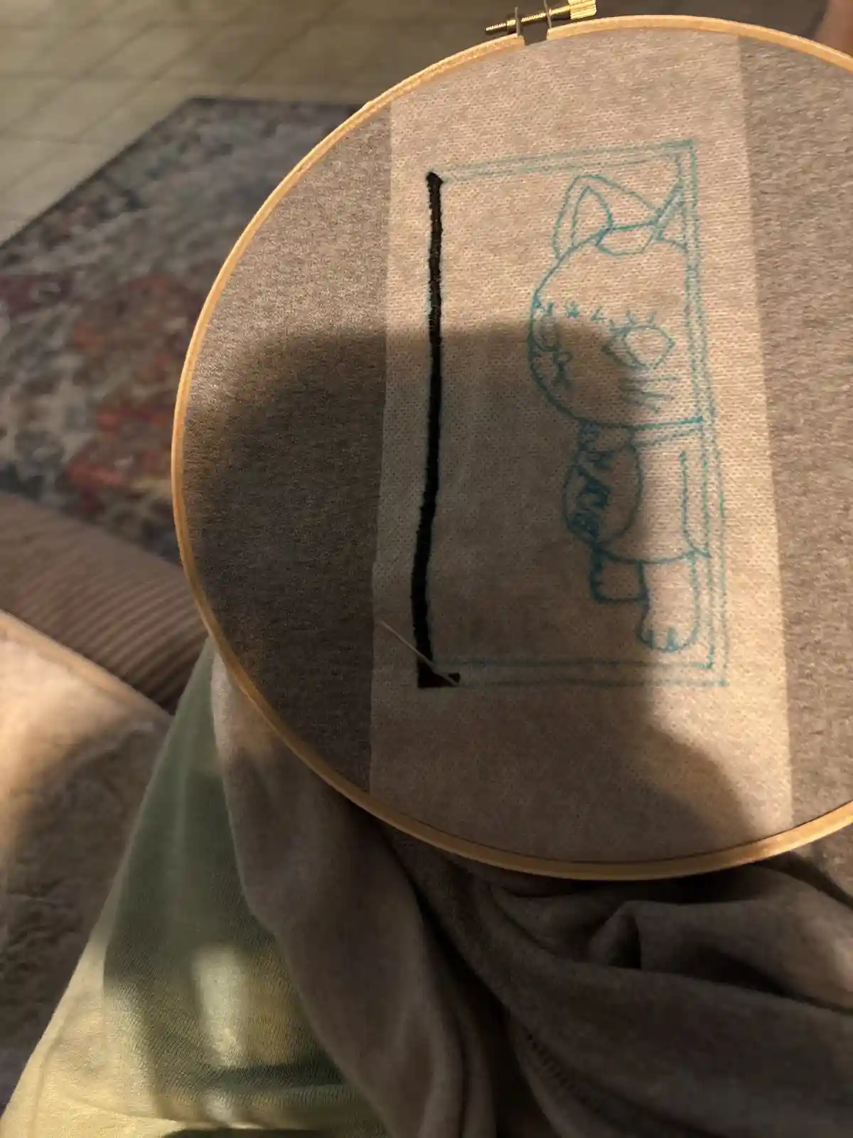 A close-up photo shows a person holding an embroidery hoop with a partially stitched design of a cartoon cat peeking through…