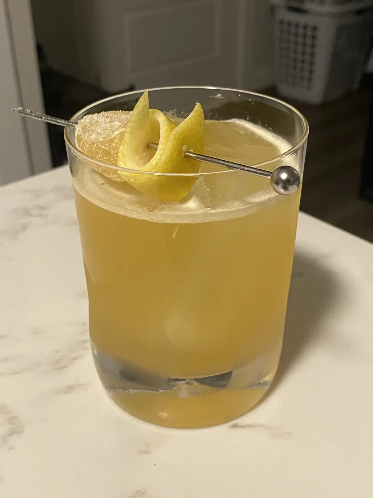 A close-up photo shows a cocktail in a rocks glass with a yellowish liquid, a lemon twist, and a sugared lemon wedge on a…