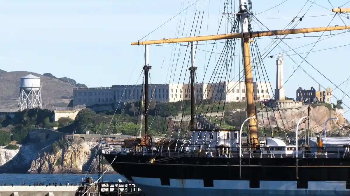 A historic sailing ship is docked in front of Alcatraz Island, with its iconic prison building and lighthouse visible in the…