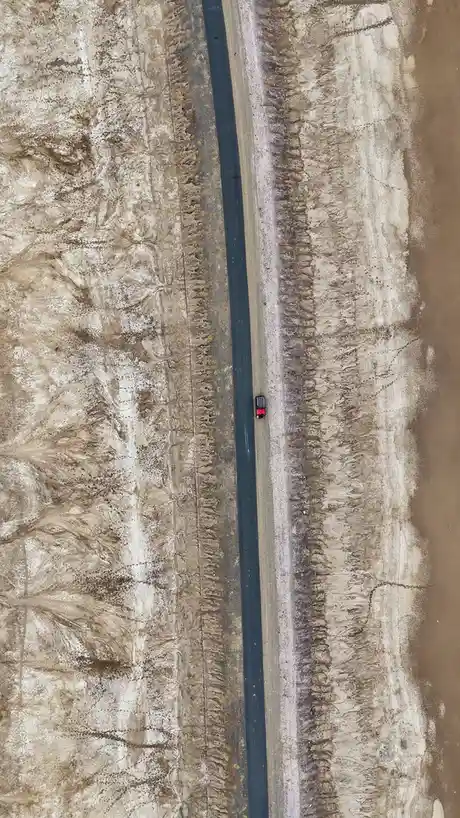An aerial view shows a single red car driving down a long, straight asphalt road through a vast, barren, and cracked desert…