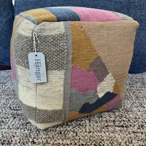 A colorful, geometric-patterned wool pouf sits on a multicolored rug, with a white tag attached that reads 'Hangar'. The…