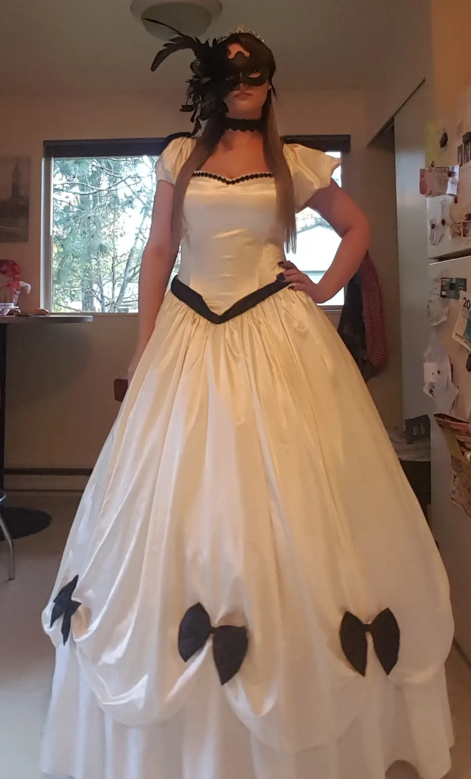 A person stands indoors wearing a large, white satin ball gown with black bows at the hem, a black lace choker, and a black…