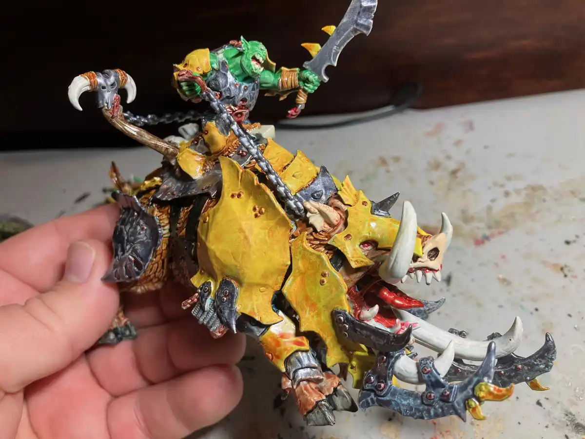 A hand holds a heavily painted miniature of a green-skinned orc riding a monstrous, spiked boar-like creature adorned with…