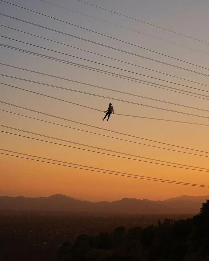 A person is silhouetted against a sunset, hanging from a power line high above a landscape with mountains and trees. The sky…