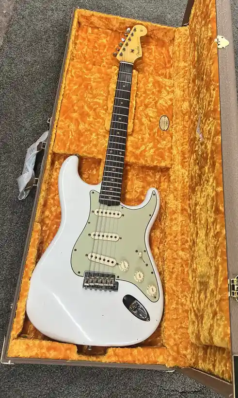 A white Fender Stratocaster electric guitar with a light green pickguard rests inside an open, orange-felt-lined hardshell…