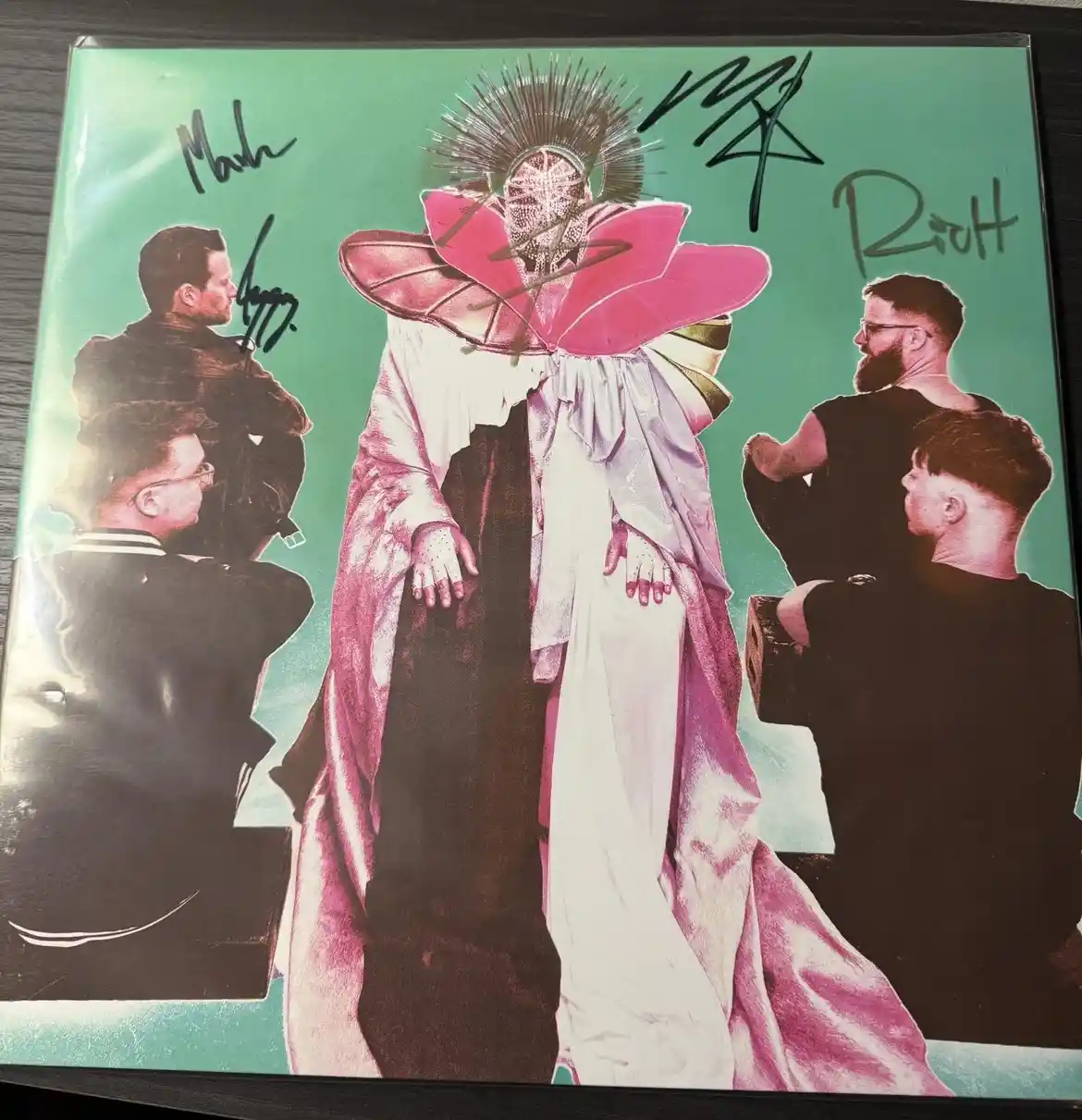 An autographed album cover featuring a central figure in a pink, flower-like headdress and flowing robe, surrounded by four…