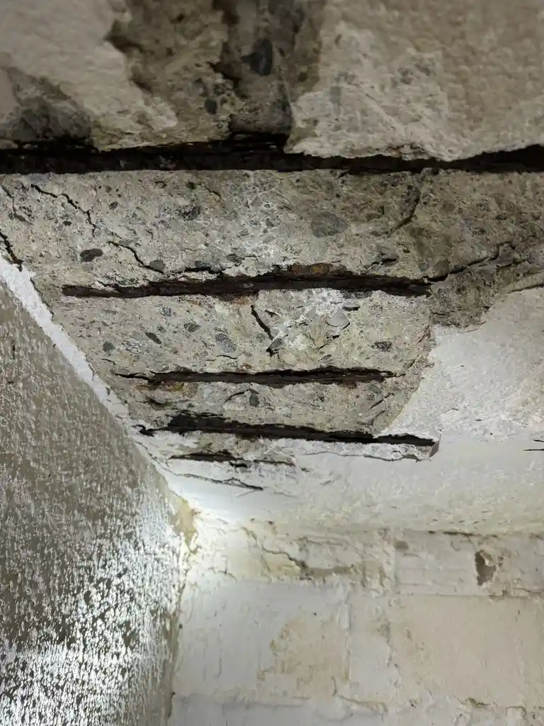 A close-up photo shows a deteriorating concrete wall corner with multiple horizontal cracks revealing dark, rusted metal…
