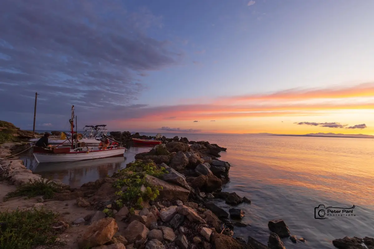 A serene sunset photograph shows fishing boats moored at a rocky shoreline, with vibrant orange and purple hues reflecting…