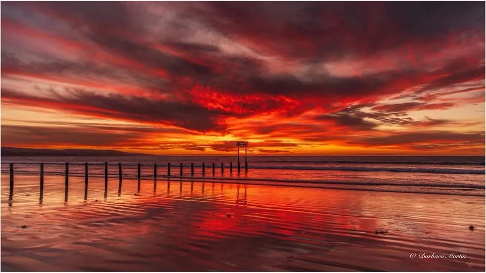 A dramatic, fiery sunset over a beach with vivid red and orange clouds reflected in wet sand, featuring a row of wooden…